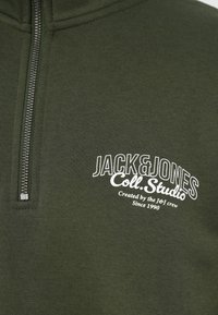 Φούτερ ελιάς με φερμουάρ στον λαιμό, κατασκευασμένο από απαλό ύφασμα, με λευκή εκτύπωση λογότυπου και κείμενο στο μπροστινό μέρος. Το φερμουάρ τρέχει κάθετα.