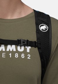 Κοντινό πλάνο ενός ατόμου που φοράει πουκάμισο χακί με λευκό κείμενο και ιμάντα μαύρου σακιδίου πλάτης που ασφαλίζει με αγκράφα στο στήθος.