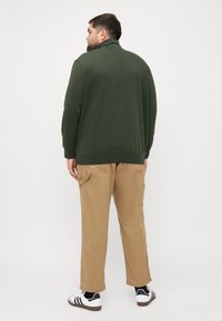 Dark green zip-up sweatshirt with a high collar, paired with light beige pants and black-and-white sneakers, viewed from the back.