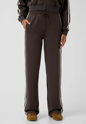 Persona che indossa pantaloni da tuta marrone scuro con strisce laterali bianche e scarpe marroni, in piedi con una mano in tasca su sfondo grigio.