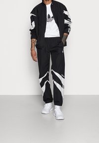 Man klädd i en svart och vit Adidas träningsdräkt och vita sneakers, stående med ena handen i fickan mot en enfärgad bakgrund.