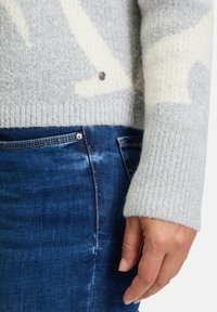 Hellgrauer Pullover mit weißem abstraktem Muster, gerippten Bündchen, einem runden silbernen Akzent und kombiniert mit dunkelblauen Jeans. Weiche Textur sichtbar.