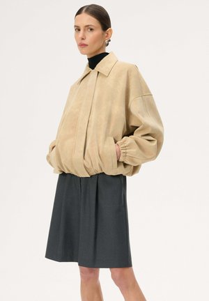 Kobieta ubrana w beżową, oversize'ową kurtkę z elastycznymi mankietami oraz ciemnoszarą spódnicę sięgającą kolan, stojąca z rękami w kieszeniach na jednolitym tle.