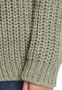 Grön stickad tröja med ett texturerat mönster; har tjockt garn och ribbade ärmslut som framhäver den kraftiga stickstilen och mjuka utseendet.