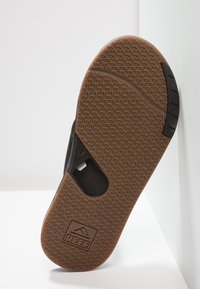 Zwarte teenslipper met een rubberen band en een bruine gestructureerde zool, met een kenmerkend ruitpatroon en logo op de hiel.