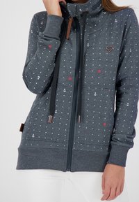 Felpa grigia con zip, decorata con pois bianchi, motivi di ancore rosse e fiori, e dettagli in pelle marrone. Presenta un colletto alto e tasche a marsupio.