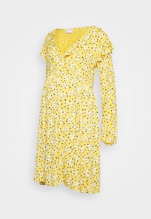 Vestido de maternidade amarelo com design de envoltório, mangas longas, decote com babados, bolinhas pretas e padrões florais brancos por todo o vestido.