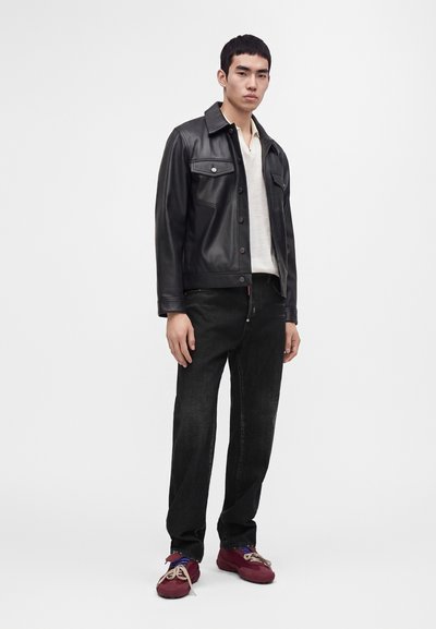 Jeune homme debout portant une veste en cuir noire, une chemise blanche, un jean noir et des baskets rouges sur un fond clair uni.