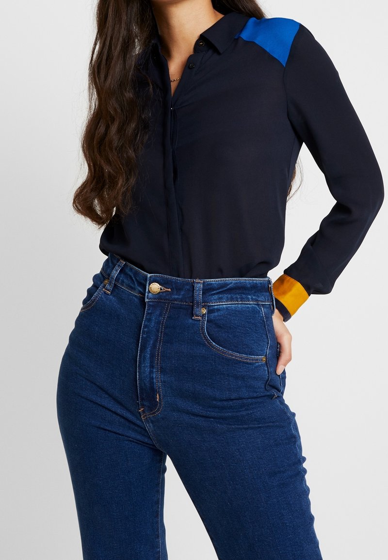 Camisa de botones navy con acento azul en los hombros, combinada con jeans oscuros de tiro alto. Tejido suave, diseño ajustado con mangas largas.