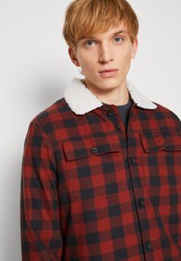 Camisa xadrez vermelho e preto com colarinho de fleece branco, botão frontal e dois bolsos no peito. Tecido de algodão com uma textura suave.