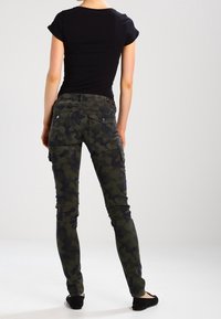 Maskáčové cargo kalhoty v tmavě zelené a černé barvě, s úzkým střihem, bočními kapsami a hladkým, odolným materiálem. Viditelný černý upnutý top.