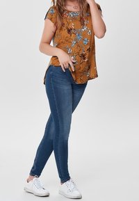 Top floral mostaza con mangas cortas y aberturas laterales, combinado con jeans ajustados azules y zapatillas blancas.