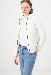 Giacca bianca trapuntata con cappuccio, caratterizzata da un motivo ondulato, chiusura con zip e bordi a contrasto; abbinata a una camicia azzurro chiaro e jeans in denim.