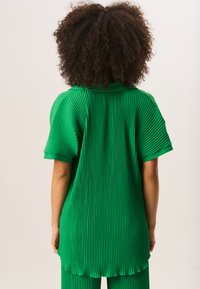 Green pleated shirt with short sleeves and a curved hem, featuring vertical ribbed texture. The collar is also visible at the back.