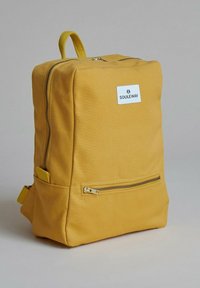 Žlutý plátěný batoh obdélníkového tvaru, s přední kapsou na zip a označením značky. Obsahuje pevné popruhy a viditelné detaily šití.