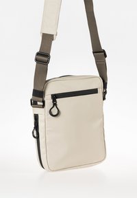Beige crossbody tas met een rechthoekige vorm, gemaakt van lichtgewicht materiaal, met zwarte ritsen, een verstelbare strap en metalen hardware accenten.