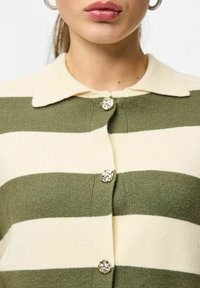 Zelena in kremasta črtasta jopica s spodnjim ovratnikom in dekorativnimi gumbi na sprednji strani; mehek tekstura in pleten material.