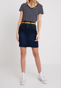 Džínová sukně v tmavě modré barvě, s předním zapínáním na knoflíky a dvěma kapsami. V kombinaci s pruhovaným námořnickým tričkem a žlutým páskem; vzhled doplňují bílé tenisky.