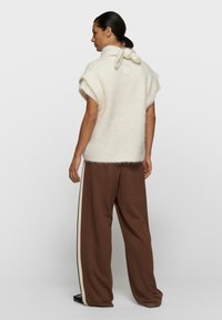 Mujer con cabello oscuro recogido en una coleta, lleva una blusa corta de manga corta color crema y borrosa con un cuello atado, y pantalones marrones sueltos con franjas laterales.