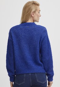 Blå stickad tröja med en texturerad yta, ribbad nederkant och ärmavslut, samt ett litet metallemblem på baksidan nära nacken.