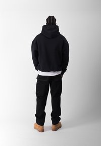 Felpa nera con un ampio cappuccio, indossata sopra una maglietta bianca, abbinata a pantaloni cargo neri e stivali da lavoro beige. Tessuto con texture, vestibilità comoda.