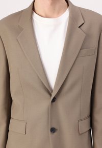 Light brown blazer with a structured design, notched lapels, two front pockets, and a single button closure. Worn over a white shirt.