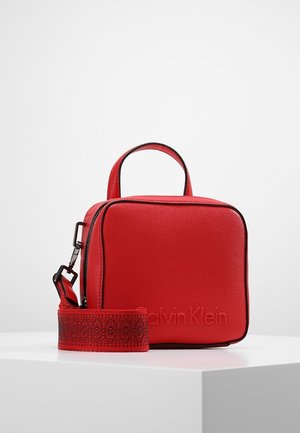 Czerwona kwadratowa torba na ramię z górnym uchwytem i wzorzystym regulowanym paskiem, z tłoczonym logo Calvin Klein z przodu, prezentowana na białej powierzchni.