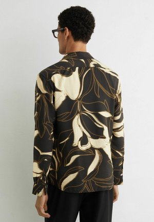 Mand iført sort skjorte med knapper og stort beige blomstermønster, set bagfra mod en ensartet lys grå baggrund.