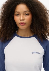 Biała bawełniana koszula z niebieskimi rękawami raglanowymi, z wyszywaniem tekstu "SUPERDRY" w granatowym na piersi. Gładka faktura, casualowy design.