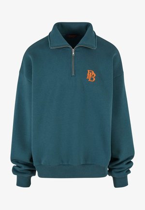 Turkusowy sweter z ćwierć zamkiem, z prążkowanymi mankietami i dołem, z pomarańczowym logo po lewej stronie klatki piersiowej. Miękka tekstura.