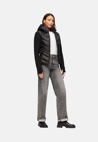 Zwarte puffer vest met capuchon, gecombineerd met een wit shirt en grijze high-waisted jeans. Donkere laarzen ronden de outfit af.