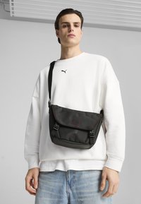 Sac messager noir avec des accents rouges, bandoulière réglable, fermeture à rabat et surface texturée arborant un logo. Porté avec un sweat-shirt blanc et un jean.