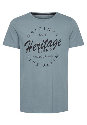 Heledy sinine puuvillane T-särk, millel on ümar kaelus, lühikesed varrukad ja must graafiline trükk, kus on tekst "Heritage Blend" ja dekoratiivsed ringid.