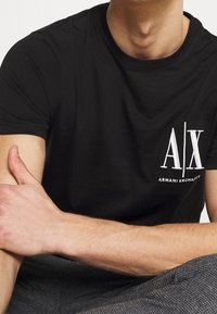 Camiseta de algodón negra con cuello redondo. Presenta un logotipo bordado en blanco "A|X" y el texto "ARMANI EXCHANGE" en el lado izquierdo.