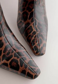 Leopard print high heels in glossy black and brown leather, featuring a pointed toe and smooth texture with visible stitching details.