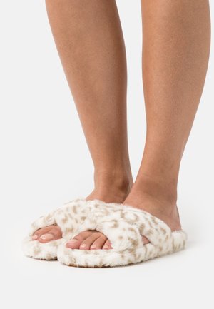 Faux-Fell Hausschuhe in Hellbeige mit gepunktetem Muster, die ein breites Banddesign und eine offene Zehenpartie aufweisen. Weiche Textur und flache Sohle.