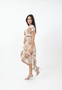 Floral Kleid in hellbeige mit Rüschensäumen und asymmetrischem Saum, das große Blumenmuster und einen gerafften Taillenbund aufweist. Klare Absatzsandalen.