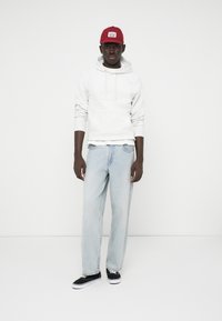 Un sweat à capuche gris, un jean bleu clair et des baskets noires avec des semelles blanches. Porte une casquette rouge avec un patch logo. Design simple et coupe décontractée.
