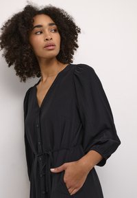 Robe noire côtelée avec décolleté en V, boutons sur le devant, manches bouffantes, poignets élastiques et ceinture à nouer. Comprend des poches latérales.