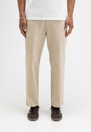 Mann trägt beige gerade geschnittene Hose, weißes Hemd und dunkelbraune Sneakers und steht mit entspannt an den Seiten hängenden Händen.