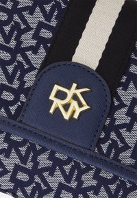 Gros plan sur un tissu bleu et blanc avec un motif répétitif du logo DKNY, une sangle rayée noire et blanche, et un patch bleu avec un emblème doré DKNY.