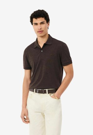 Joven con una camisa polo de manga corta de color marrón oscuro metida en pantalones crema claros con un cinturón negro, de pie frente a un fondo liso.