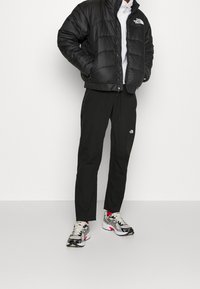 Osoba ubrana w czarną kurtkę puchową North Face, czarne spodnie North Face oraz białe, czarne i czerwone buty sportowe stoi na białej podłodze.