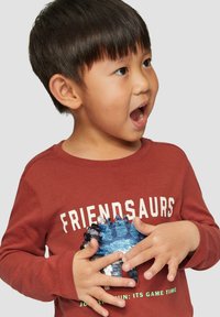 Rode langharige shirt met de tekst "FRIENDSAURS" en een blauwe sequin dino-ontwerp. Glad materiaal en standaard pasvorm, gedragen door een kind.