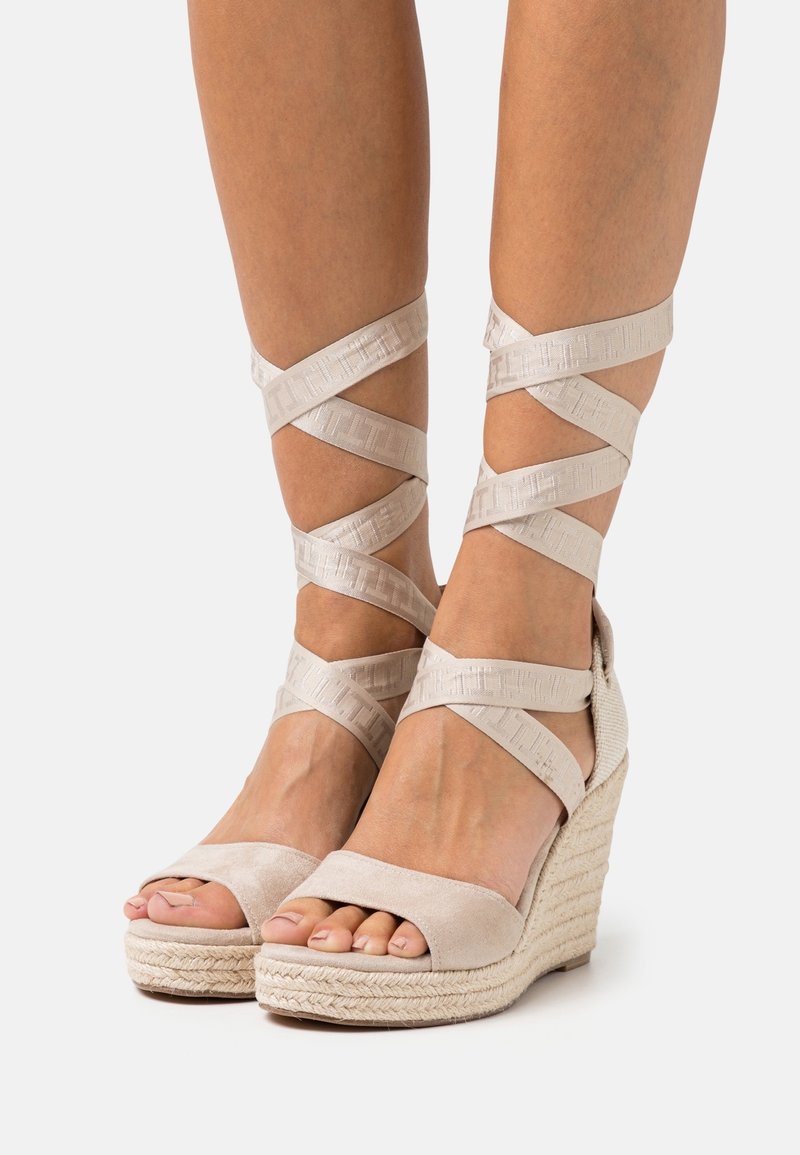 Pieds portant des sandales compensées beiges avec semelles en corde tressée et brides croisées à la cheville sur un fond clair uni.