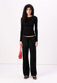 Crna dugih rukava majica, široke crne hlače, crvena torba, i obuća s leopard uzorkom. Model stoji nasuprot jednostavne pozadine.