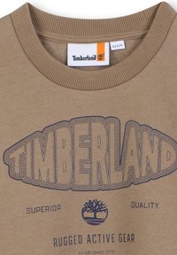 Felpa in cotone beige con stampa blu con la scritta "TIMBERLAND", logo dell'albero e le scritte "SUPERIOR", "QUALITY" e "RUGGED ACTIVE GEAR."