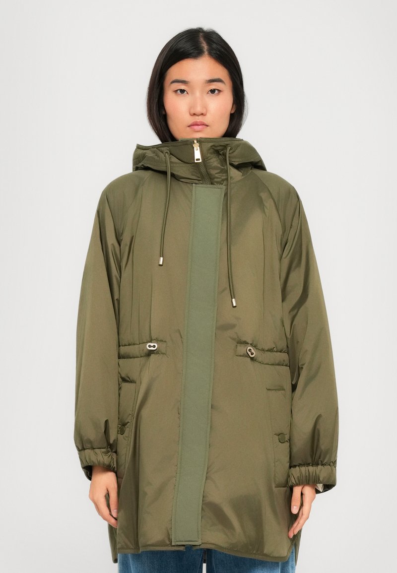 Woman wearing an oversized olive green hooded parka with drawstrings, zip closure, and elastic cuffs, standing against a plain background.