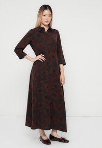 Femme debout avec une main sur la hanche, portant une robe foncée à manches longues et boutons, ornée de grands motifs floraux rouges, avec des ballerines bordeaux.
