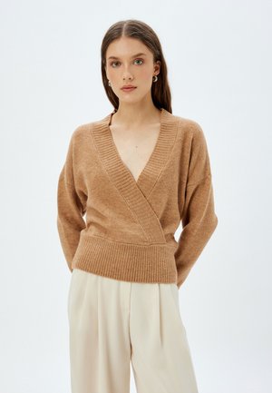 Maglione lavorato a maglia color beige con scollatura a V profonda, polsini e orlo a costine. Vestibilità rilassata con spalle scese, abbinato a pantaloni larghi color crema.